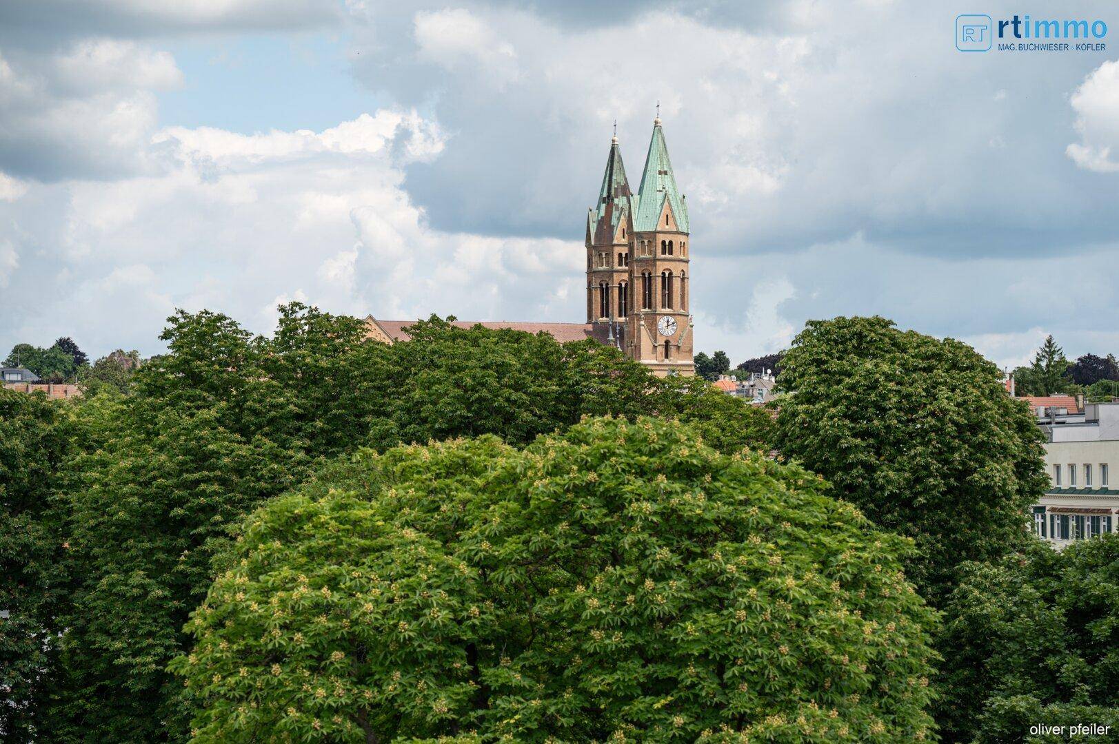 Malerischer Blick auf eine Kirche und grüne Baumwipfel unter blauem Himmel.
