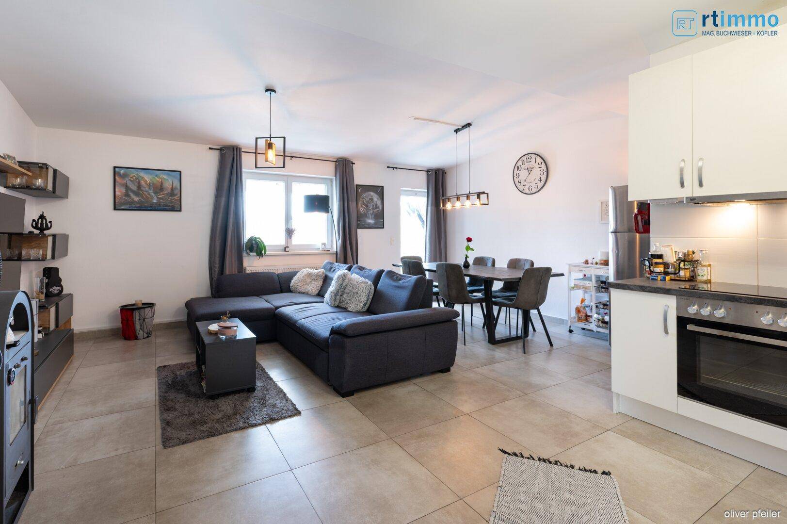 Offener Wohn-Essbereich mit moderner Küche, dunklem Sofa und großem Esstisch auf Fliesenboden.
