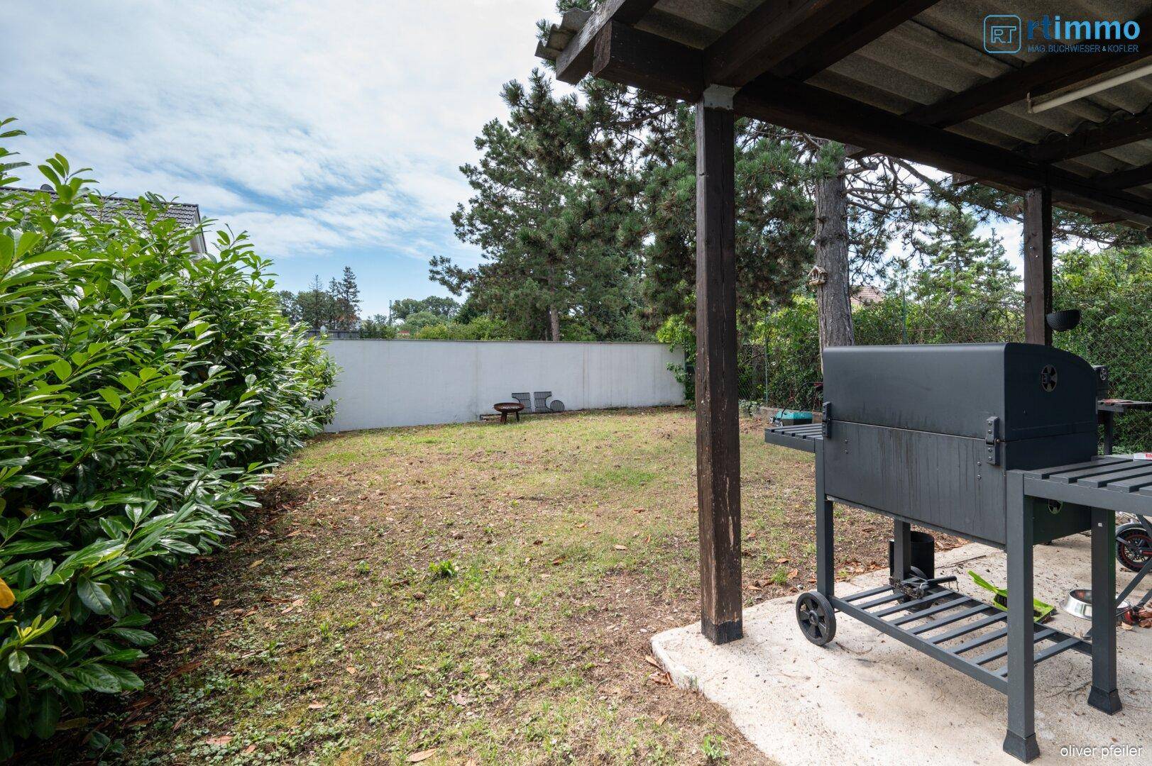 Großer Gartenbereich mit Rasenfläche, Grillplatz unter einer Überdachung und hohen Hecken.