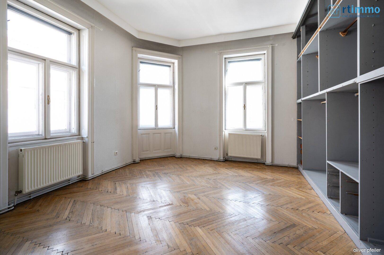 Geräumiges Zimmer mit Fischgrätparkett, großen Fenstern und einem praktischen Einbauregal.