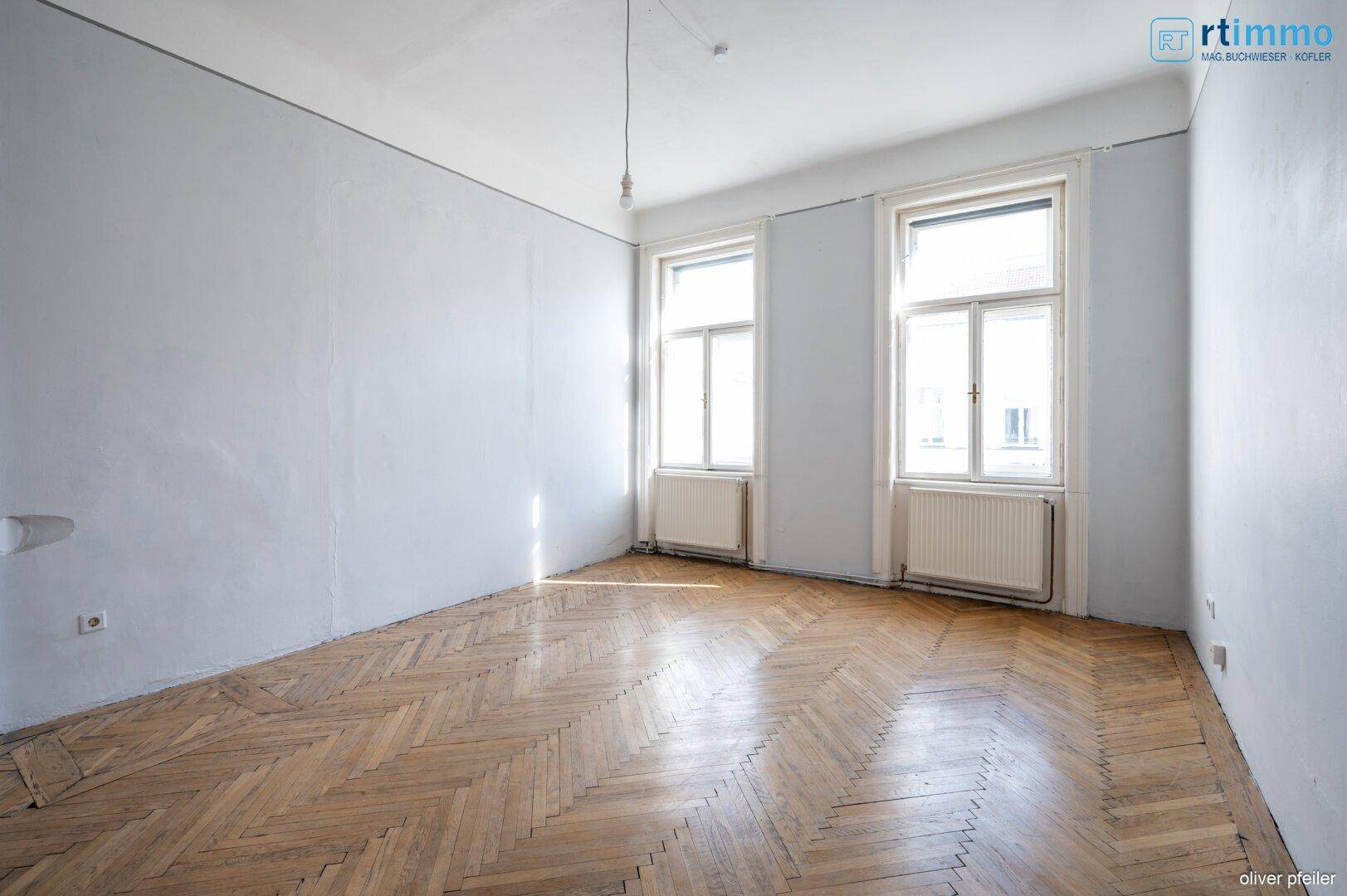 Helles Zimmer mit Fischgrätparkett und großen Fenstern, das viel natürliches Licht hereinlässt.