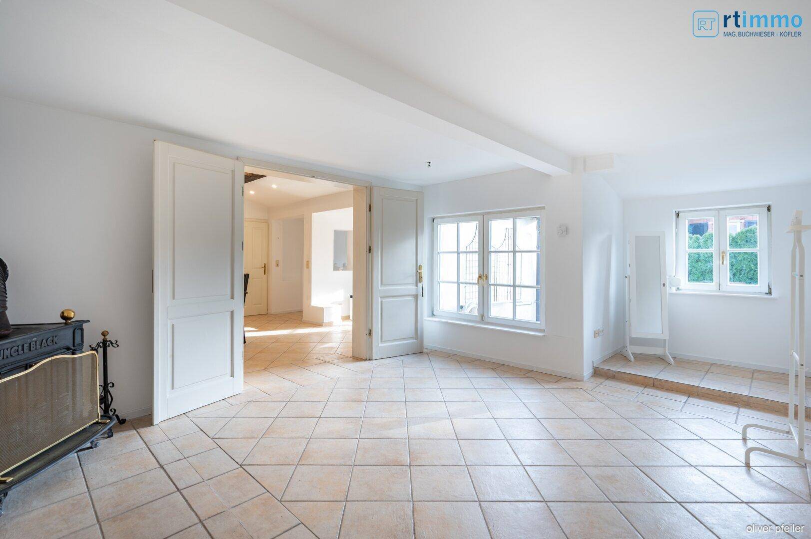 Helles Wohnzimmer mit Fliesenboden, großen Fenstern und Blick in den grünen Gartenbereich.