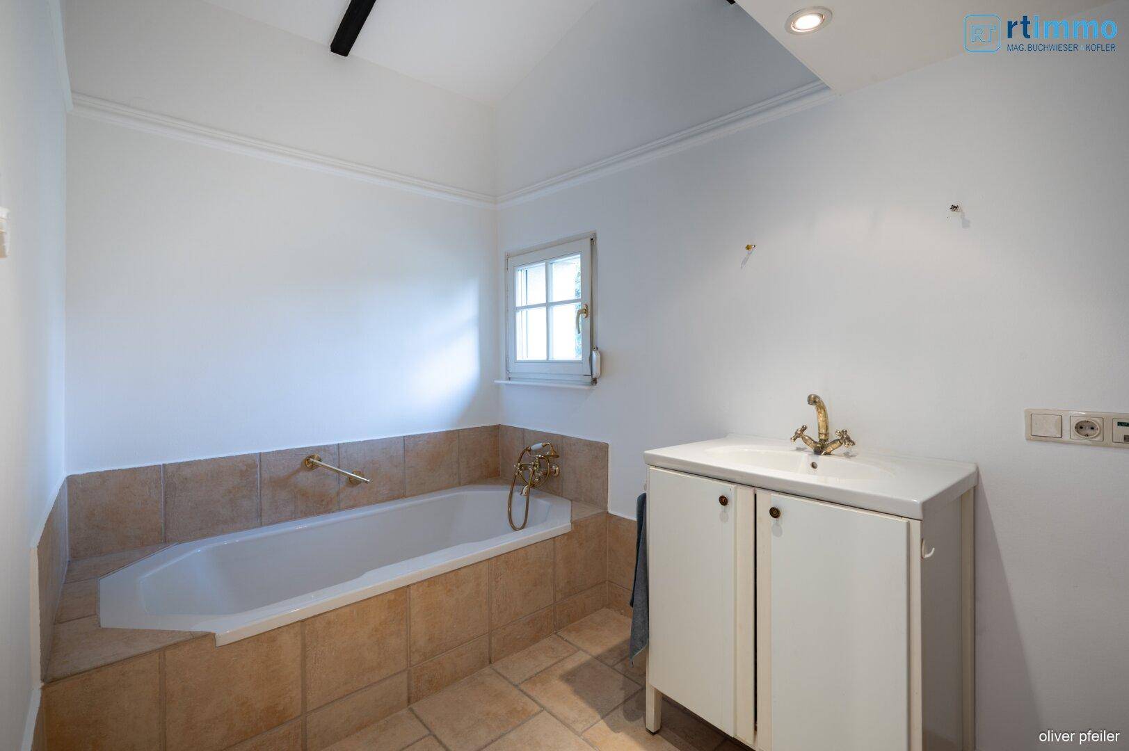 Helles Badezimmer mit Badewanne, Fenster und beigefarbenen Fliesen an Wand und Boden.