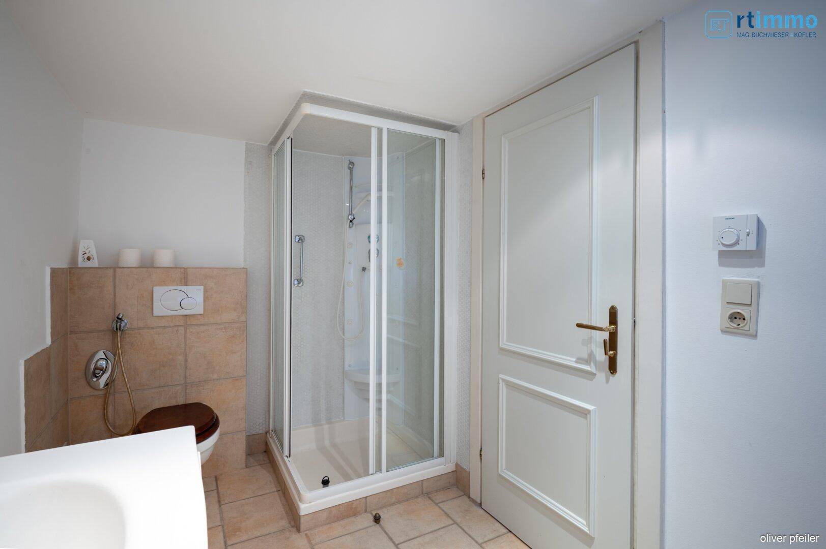 Badezimmer mit Duschkabine, wandhängendem WC und beigefarbenen Wand- und Bodenfliesen.