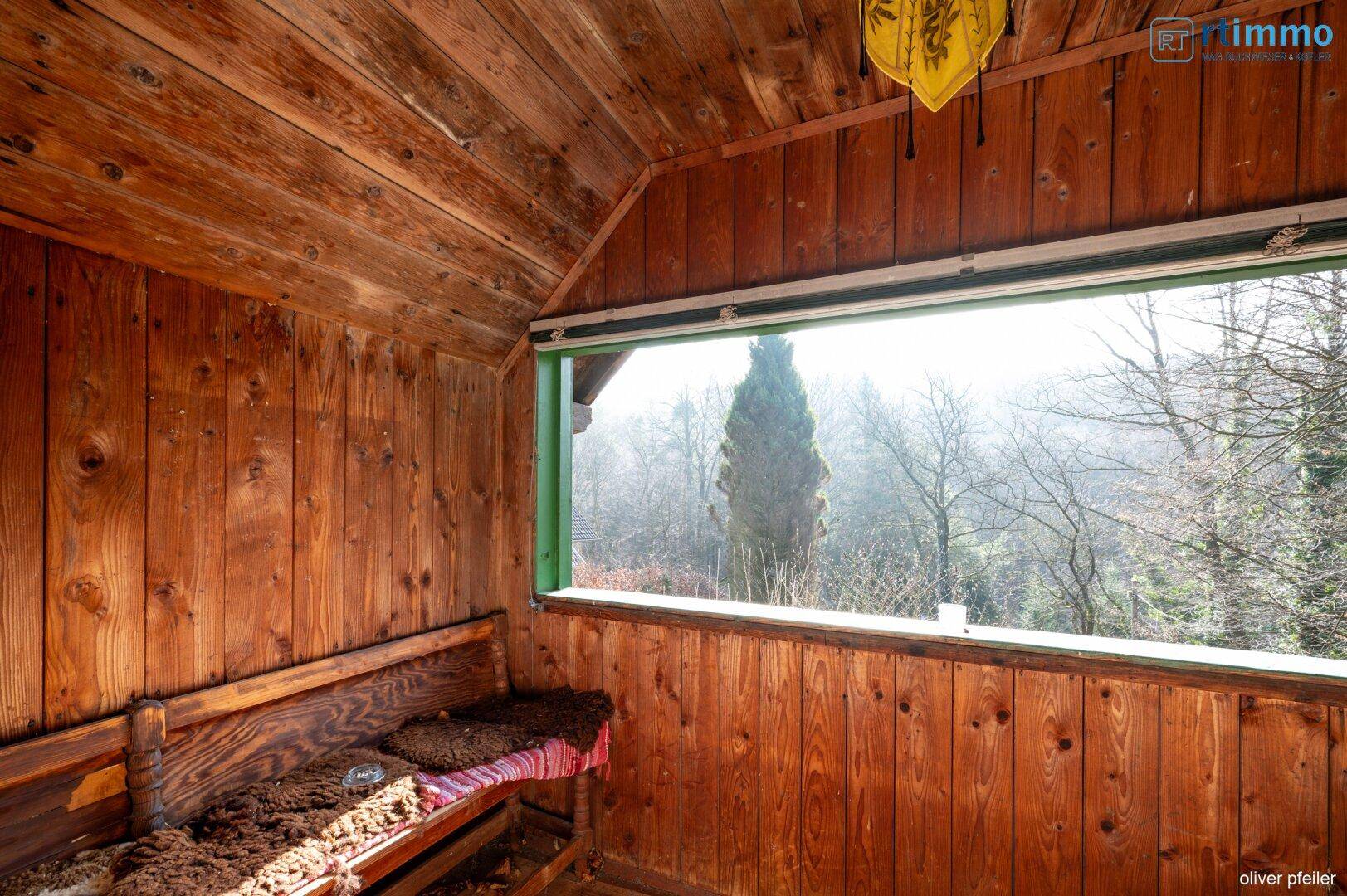 Gemütlicher, holzvertäfelter Raum mit breitem Fenster und Blick ins Grüne, ideal zum Verweilen.