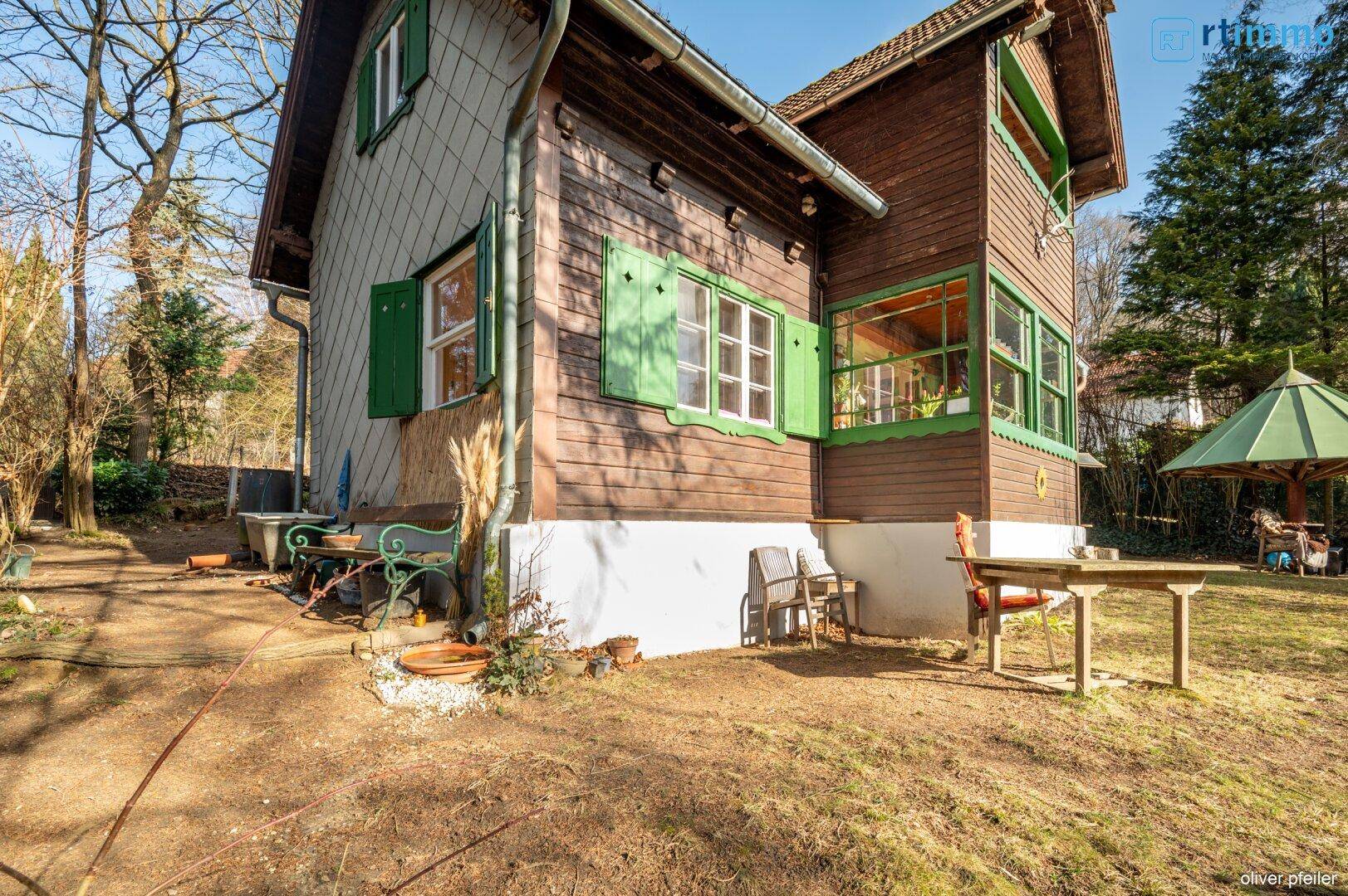 Seitenansicht des Holzhauses mit grünen Fensterläden und einem kleinen Sitzbereich im Garten.