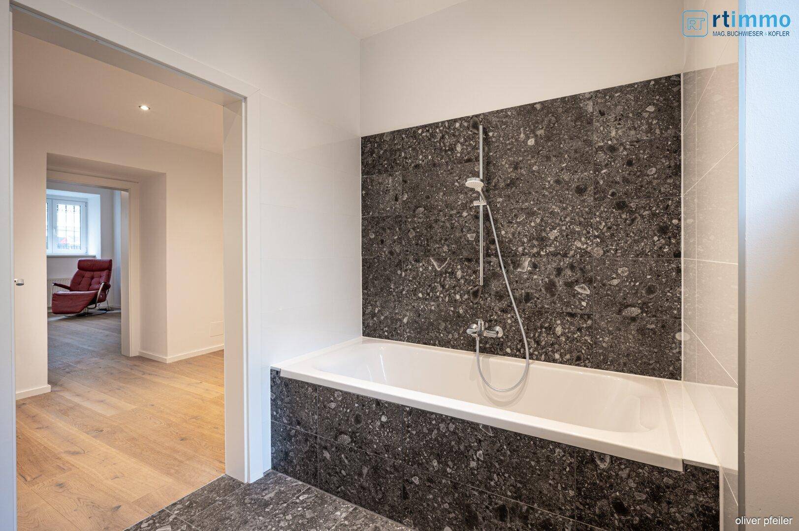 Badezimmer mit dunklen Fliesen und Badewanne, mit Blick in den angrenzenden Wohnbereich.