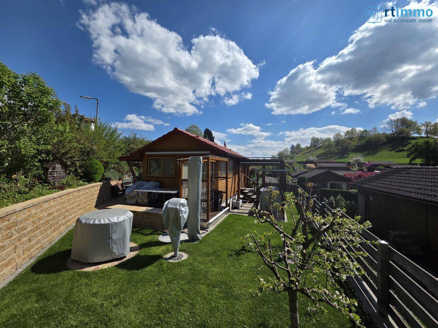 Sonniger Garten mit Rasenfläche, Steinmauer und einem Holzhaus, umgeben von grüner Natur.