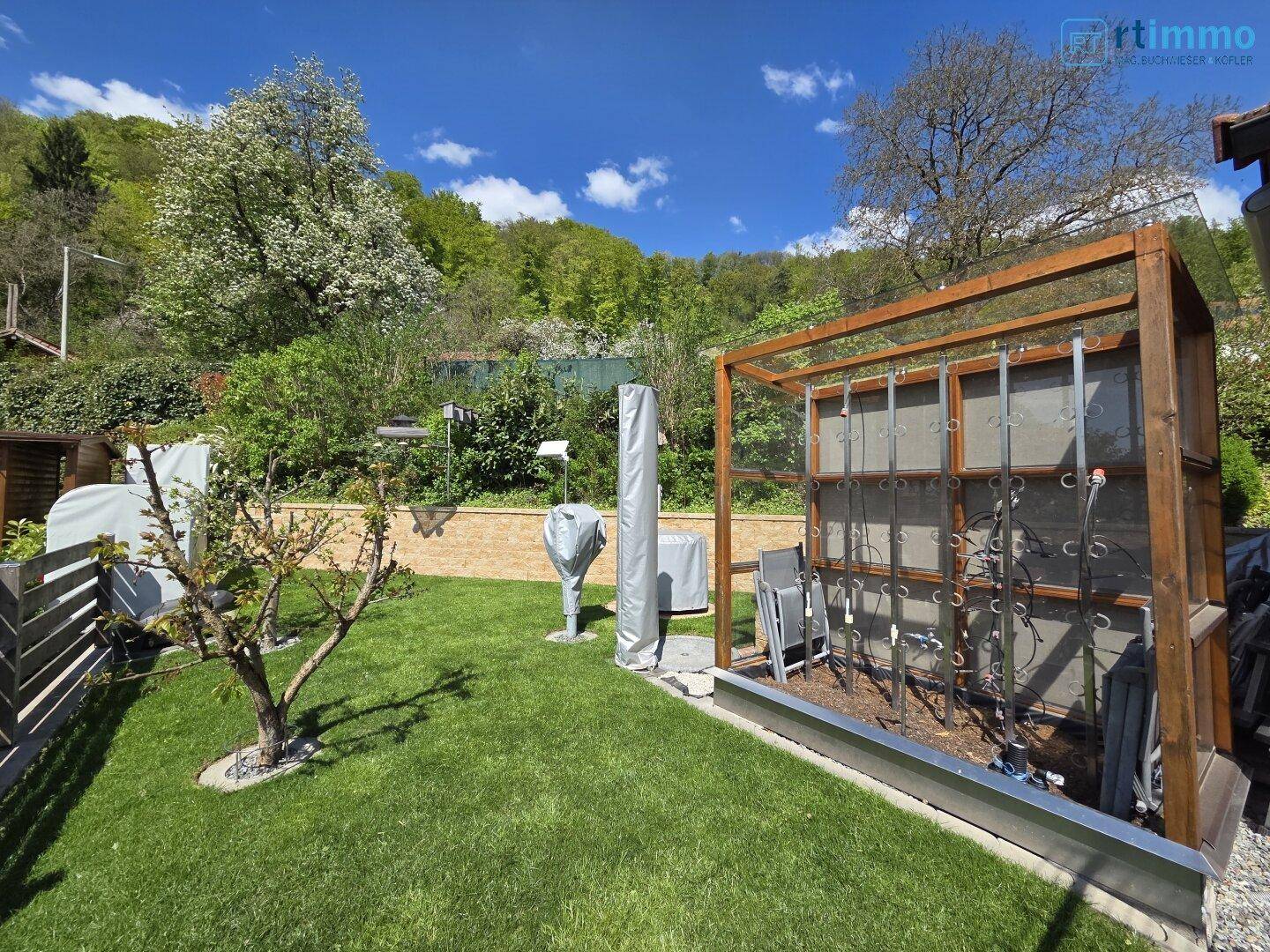 Gartenbereich mit einer Holzkonstruktion, die als Rankhilfe oder für Gartengeräte dient, und einer Steinmauer.