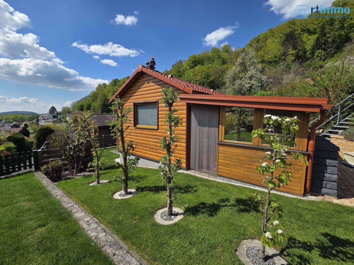 Gepflegter Garten mit blühenden Obstbäumen und einem Holzhaus unter strahlend blauem Himmel.