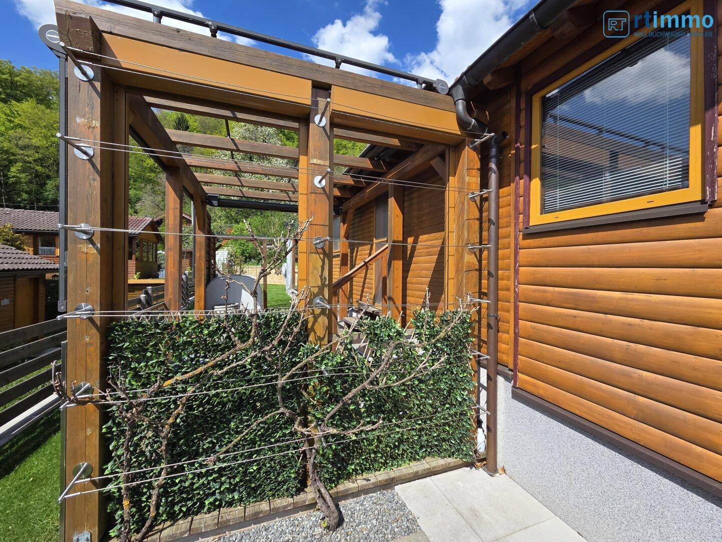 Detailansicht der Holzfassade des Hauses mit einer begrünten Pergola-Konstruktion und Kiesweg.