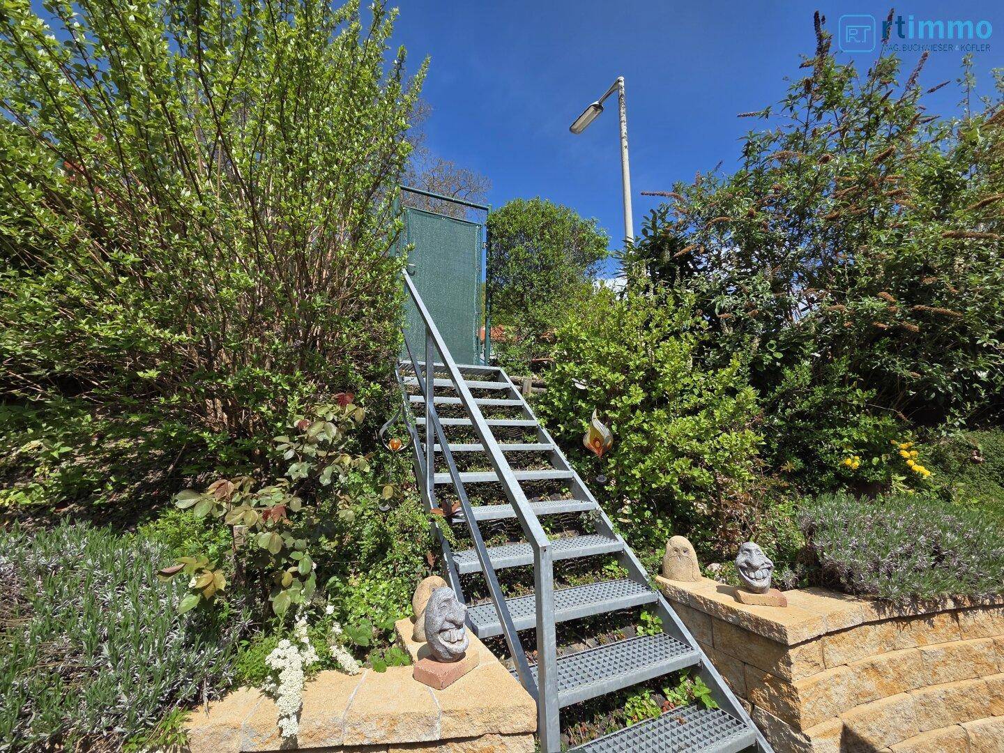 Metalltreppe im Garten, umgeben von üppiger Vegetation und einer Steinmauer.