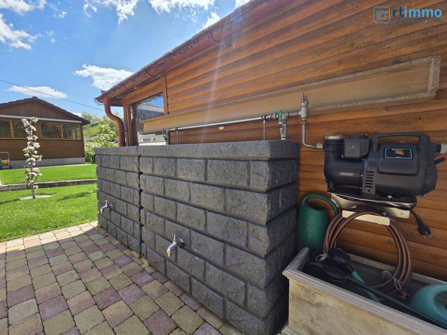 Außenbereich des Holzhauses mit Regenwassersammelbehältern und einer Wasserpumpe für den Garten.