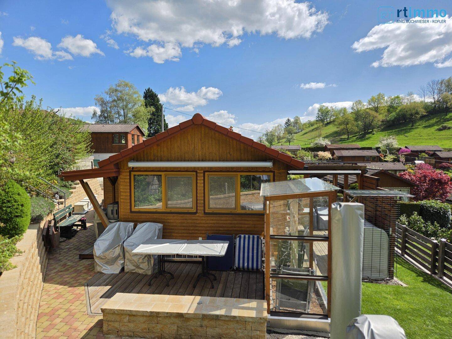 Sonniger Terrassenbereich mit Gartenmöbeln und einem kleinen Gewächshaus neben dem Holzhaus.