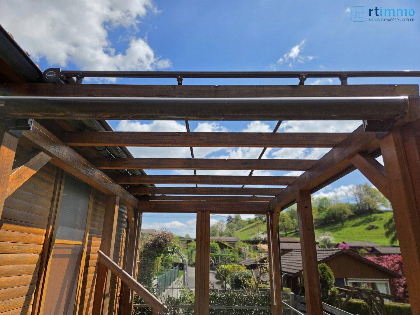 Überdachte Holzpergola mit Blick auf die umliegende grüne Landschaft und die Dächer der Häuser.