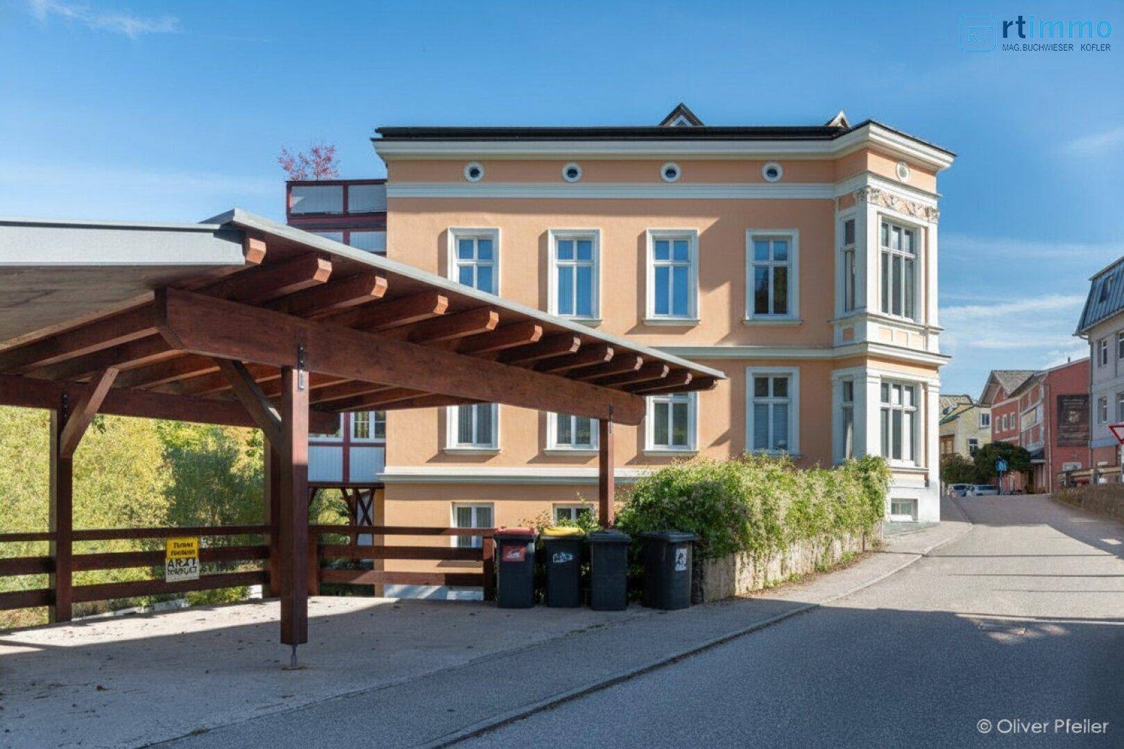 Historisches Gebäude mit Carport und Mülltonnen am Straßenrand.