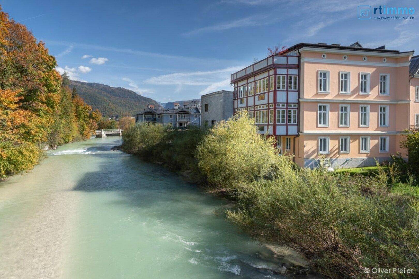 Historisches Gebäude am Fluss mit Herbstbäumen und Bergen im Hintergrund.