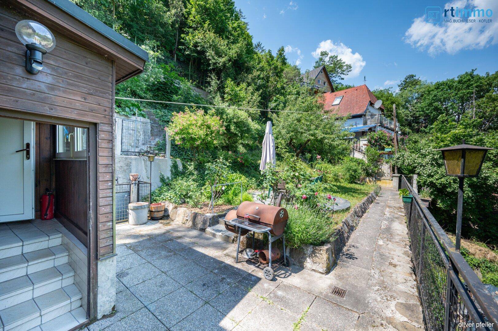 Gepflasterte Terrasse mit Grillbereich und Blick auf den bewaldeten Hang.