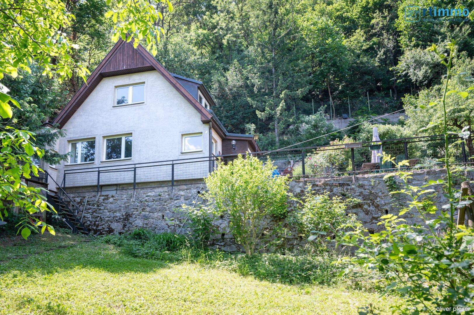 Hausansicht von unten mit großem Garten und bewaldetem Hang im Hintergrund.