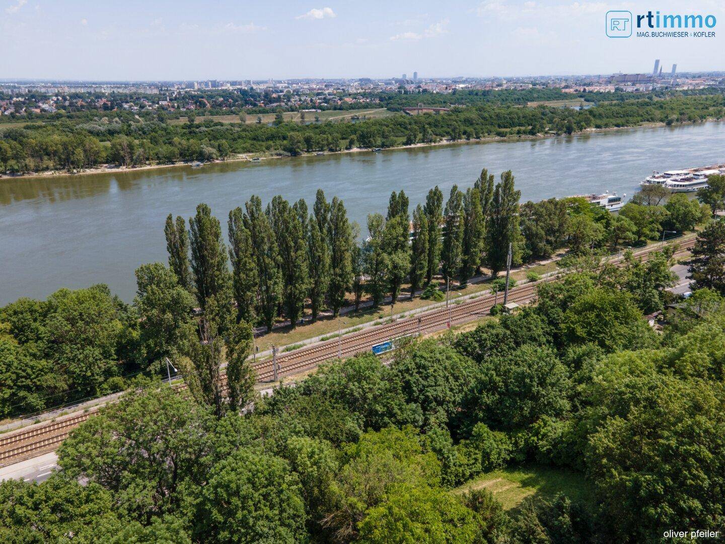 Panoramablick auf einen breiten Fluss, Bahngleise und die grüne Landschaft mit Stadt in der Ferne.