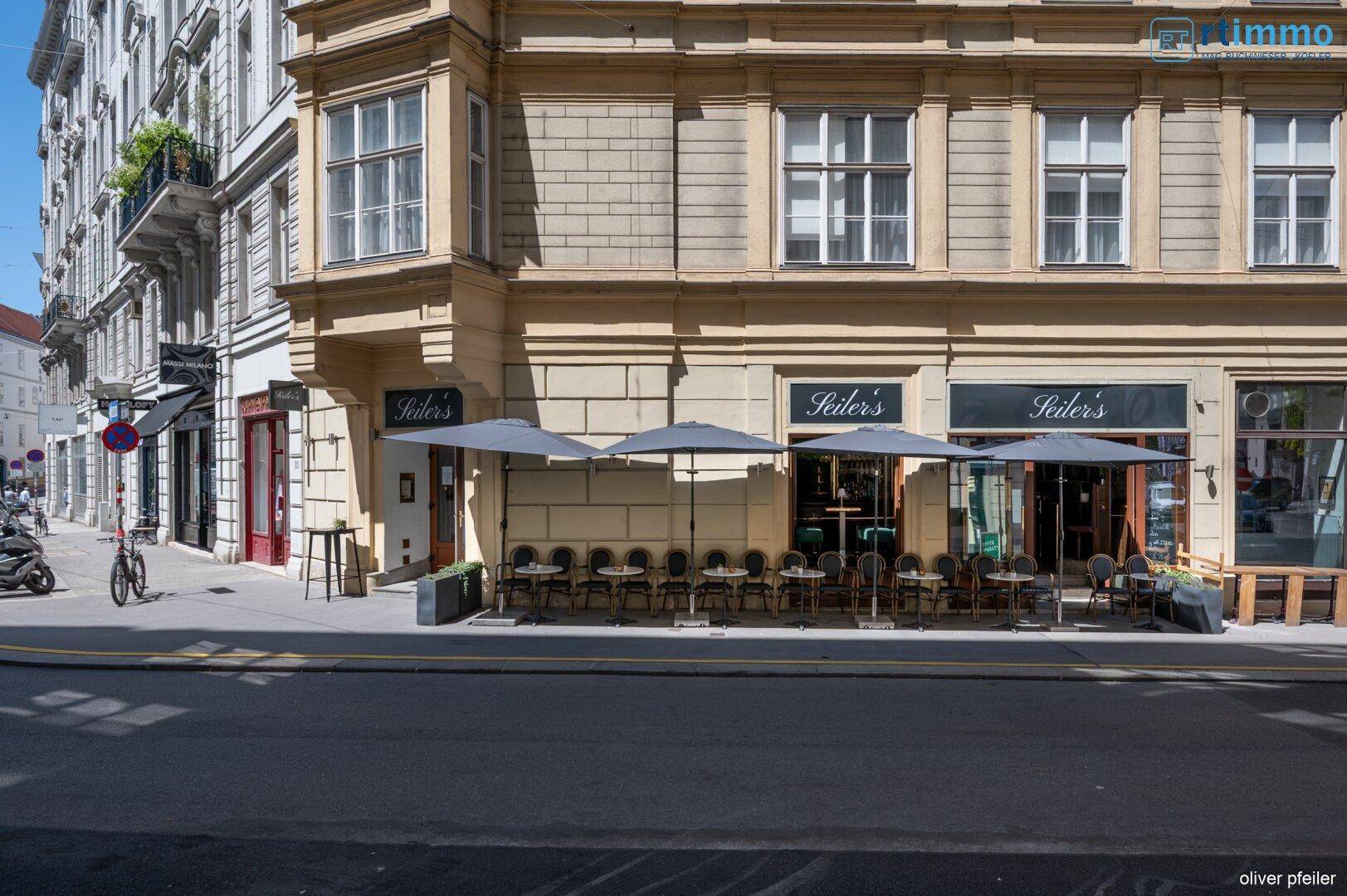 Ansicht eines historischen Gebäudes mit einer Außenterrasse und Sonnenschirmen an einer belebten Straße.