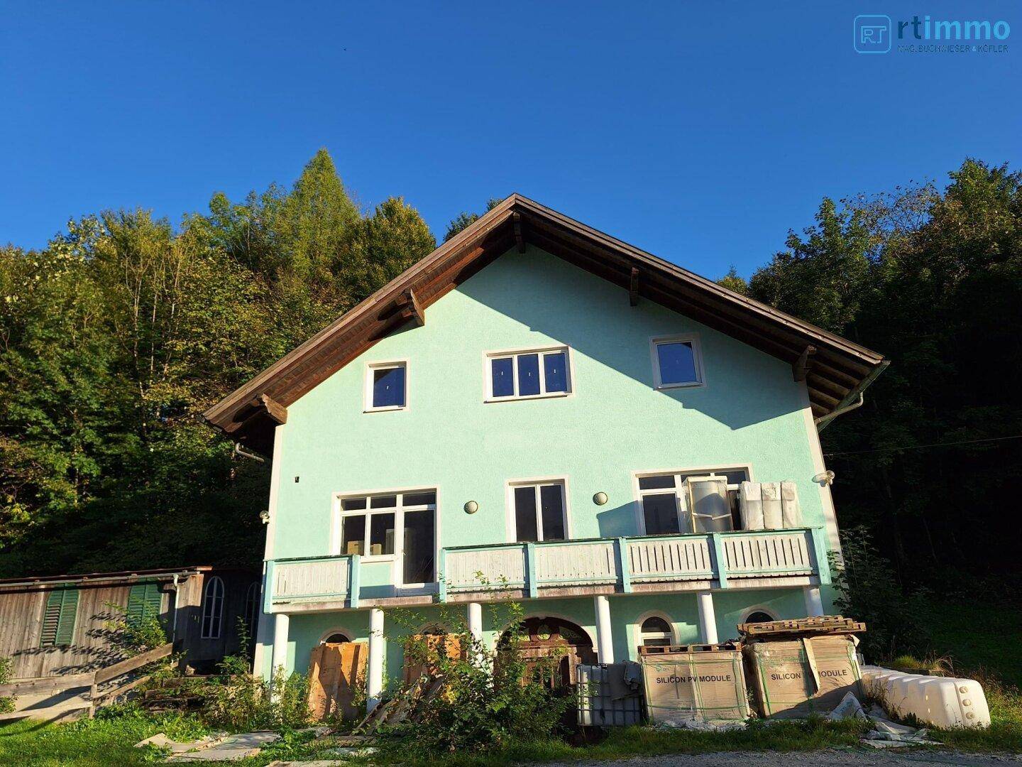 Freistehendes Haus mit hellgrüner Fassade und Holzbalkonen vor bewaldetem Hügel.
