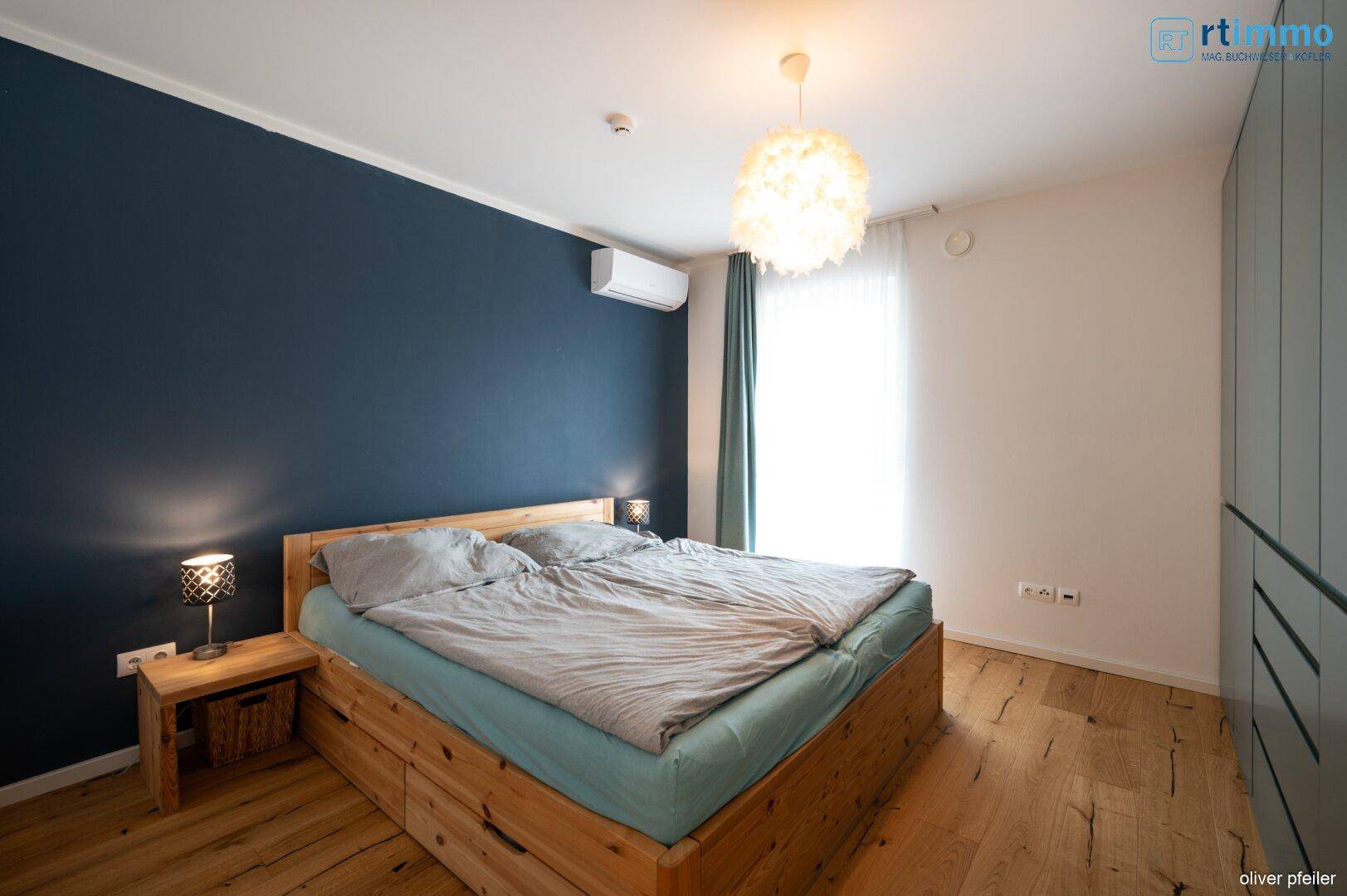 Schlafzimmer mit Holzbett, blauer Akzentwand und Klimaanlage, hell und modern eingerichtet.