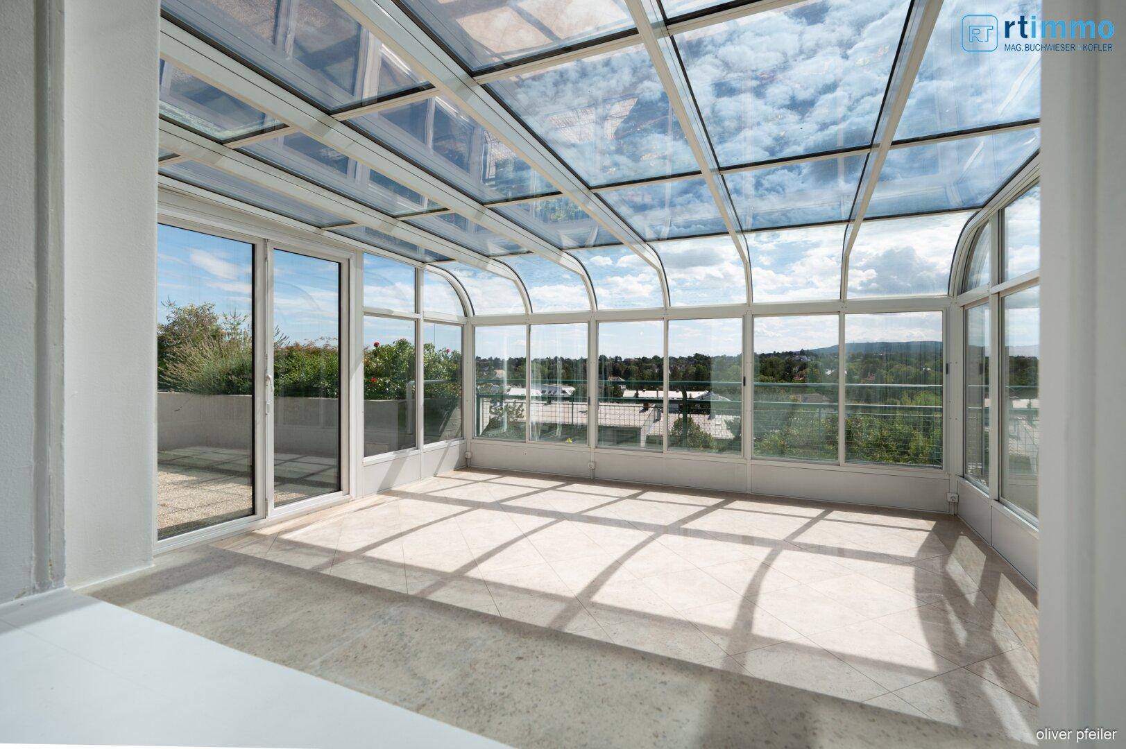 Sonniger Wintergarten mit Glasdach, Fliesenboden und Panoramablick auf die Umgebung.