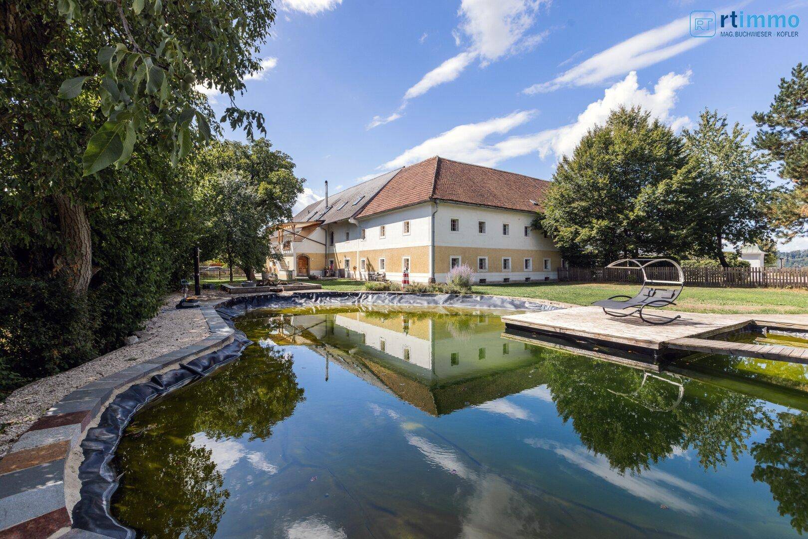 Blick auf die historische Fassade des Gebäudes mit einem großen Teich und einer Holzplattform im Garten.