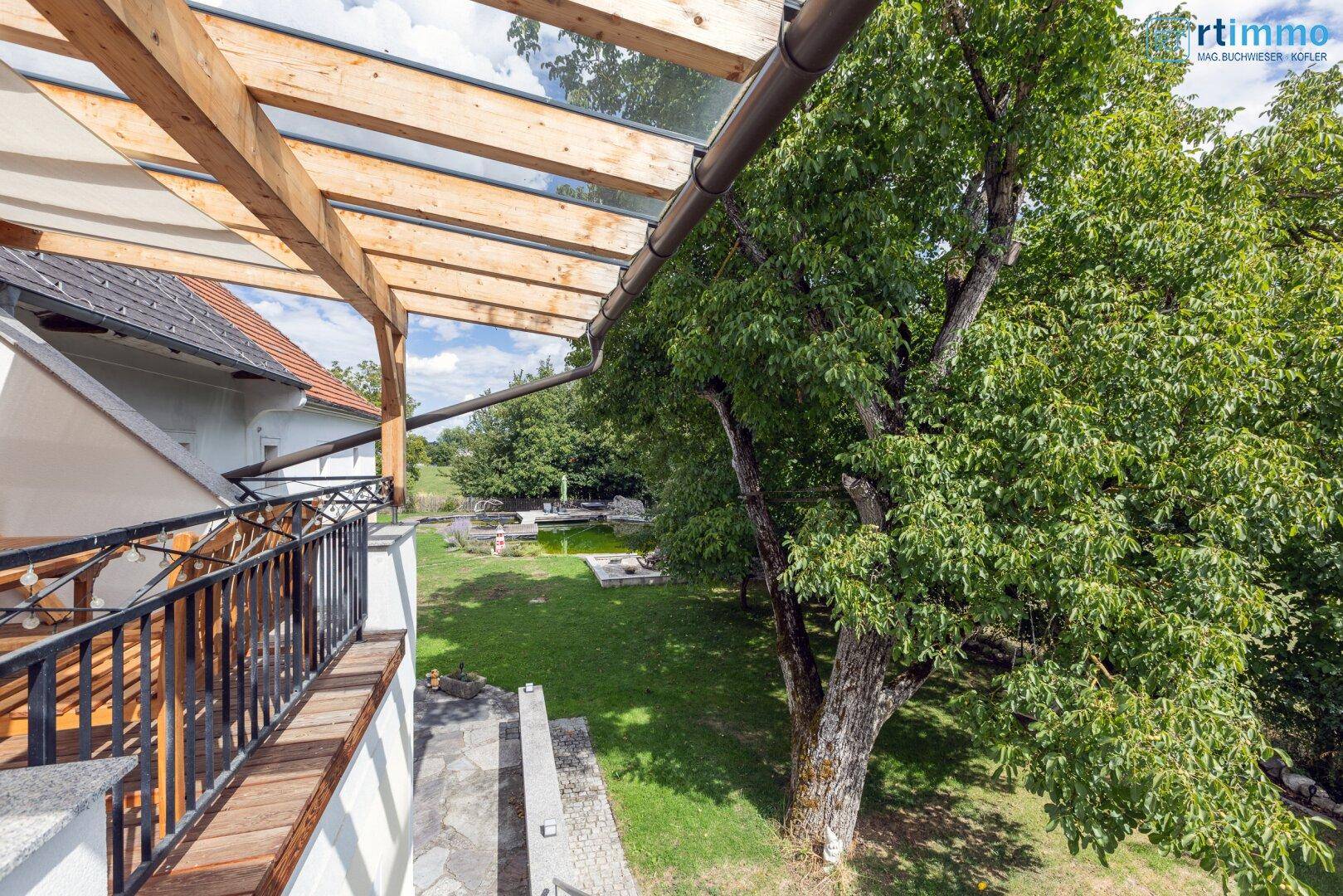 Blick von der überdachten Terrasse auf den weitläufigen Garten mit Teich und alten Bäumen.