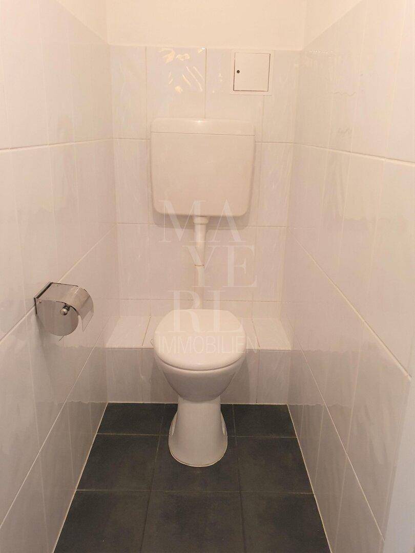 Separates WC mit weißen Fliesen an Wänden und dunklem Fliesenboden, in gutem Zustand.