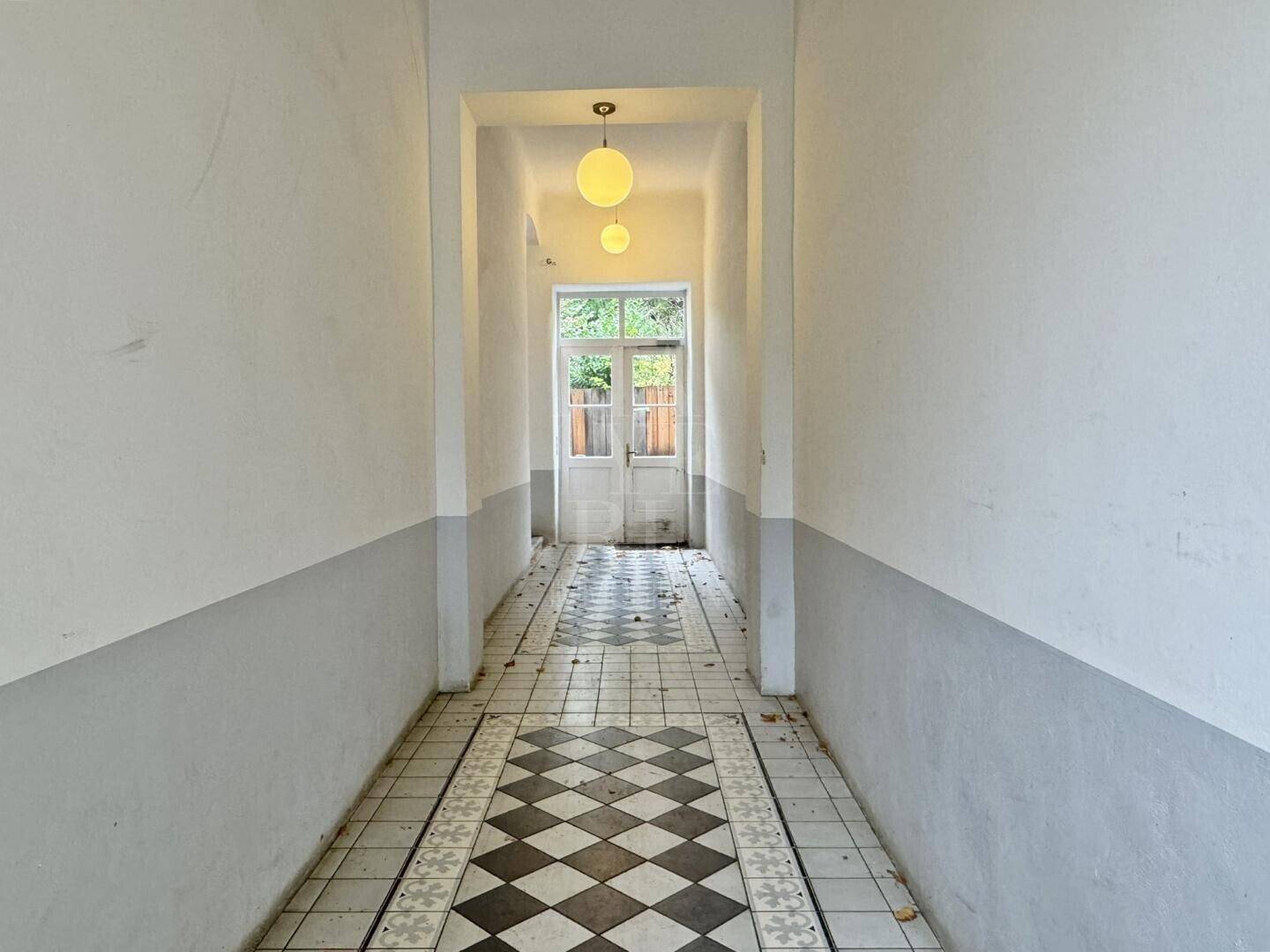 Langer, schmaler Flur mit auffälligem schwarz-weiß gemustertem Fliesenboden und einer Tür am Ende, die ins Freie führt.