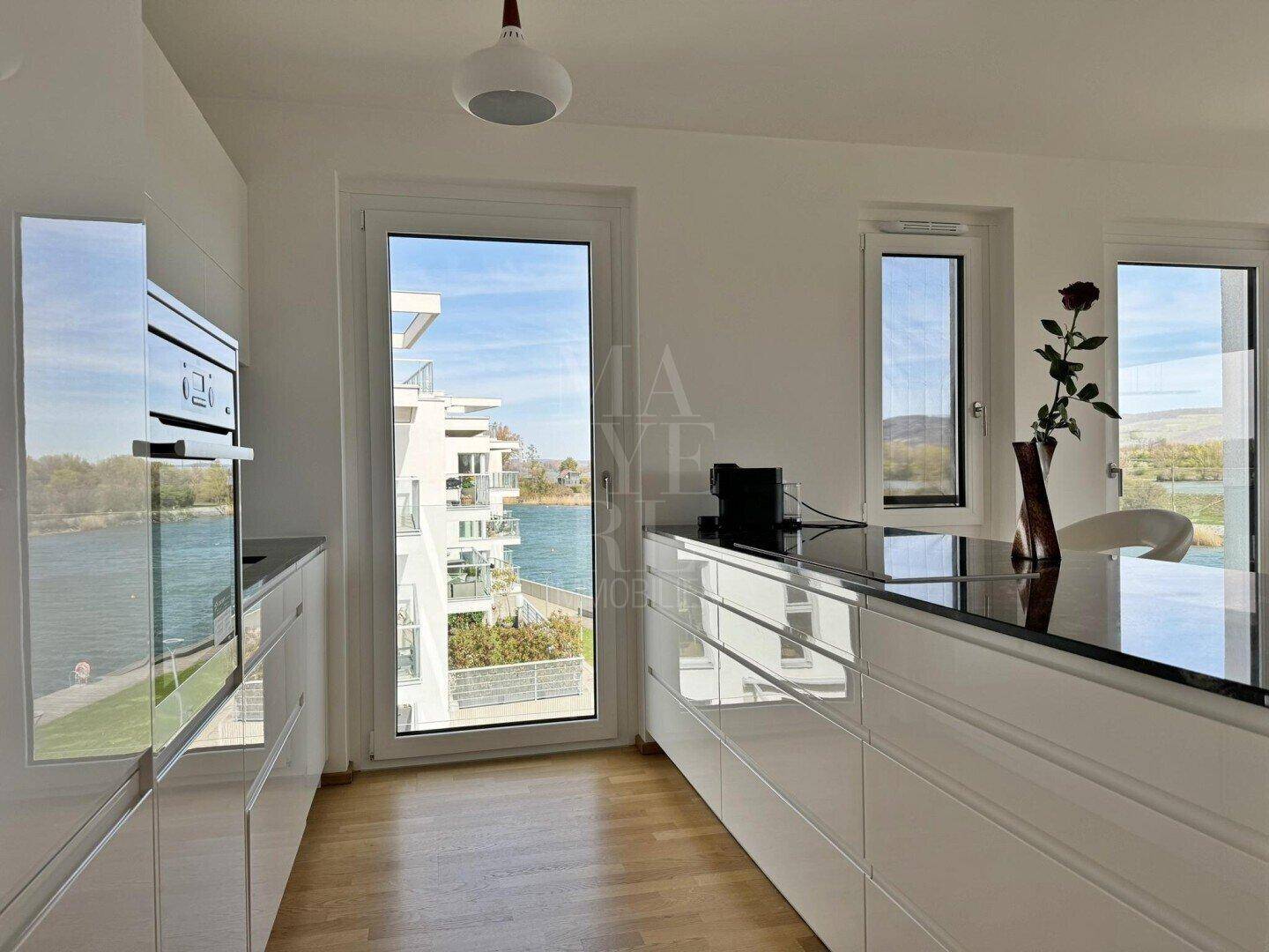 Moderne Küche mit weißen Hochglanzfronten, dunkler Arbeitsplatte und Seeblick.