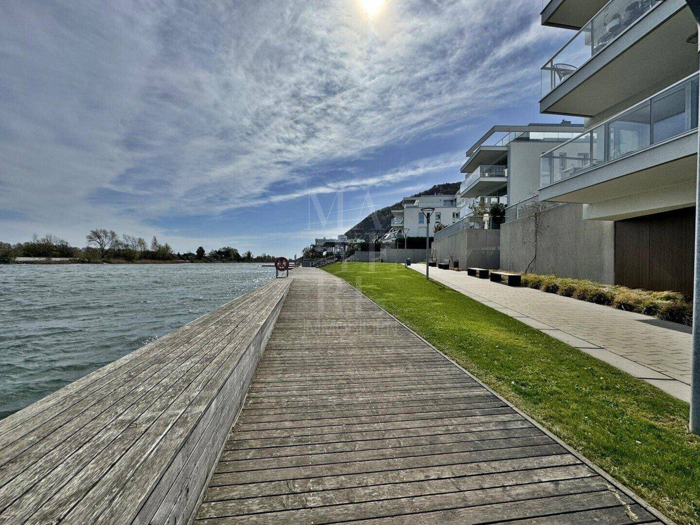 Promenade am Seeufer mit Holzsteg und modernen Wohngebäuden im Hintergrund.