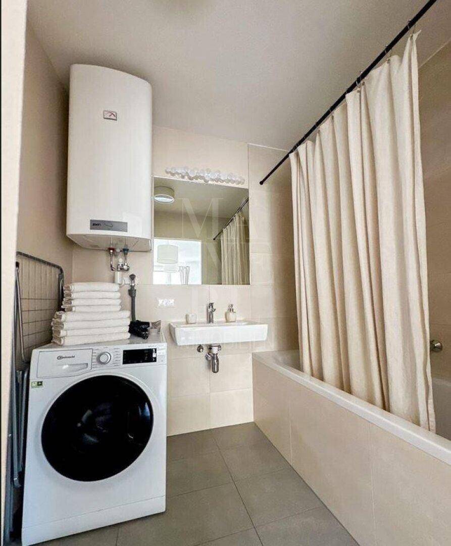 Helles Badezimmer mit Badewanne, Duschvorhang, Waschbecken und integrierter Waschmaschine.