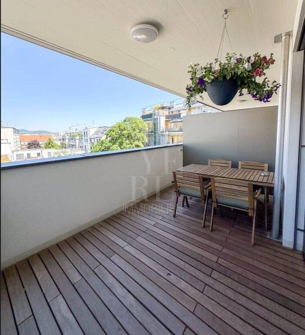 Sonniger Balkon mit Holzboden und Holzmöbeln, bietet einen weiten Blick über die Stadtlandschaft.