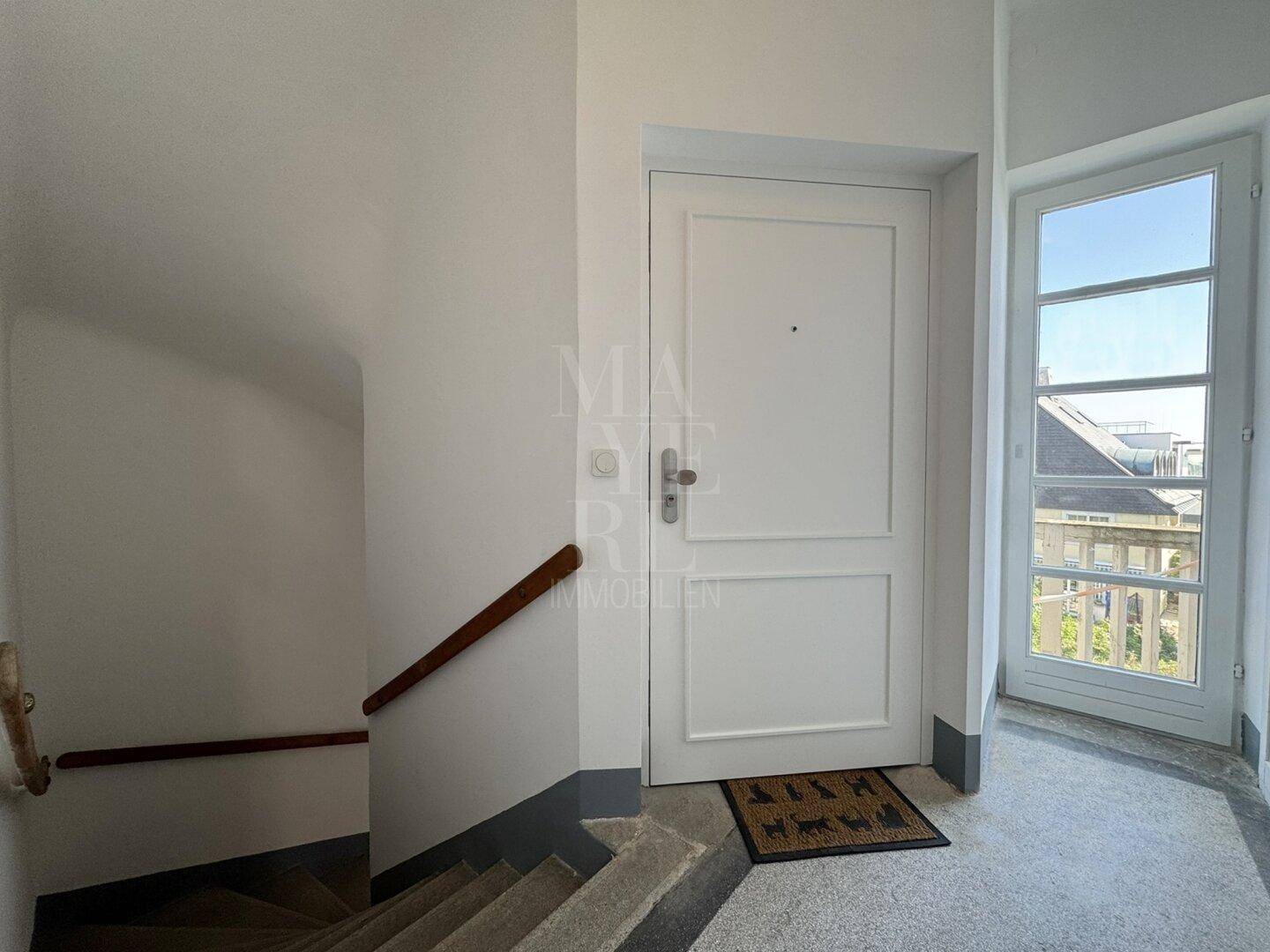 Eingangsbereich mit weißer Tür, Betontreppe und Fenster, das Tageslicht hereinlässt.