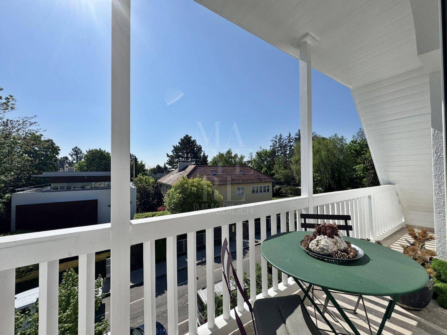 Sonniger Balkon mit weißen Holzgeländern und Sitzgelegenheiten, umgeben von viel Grün.