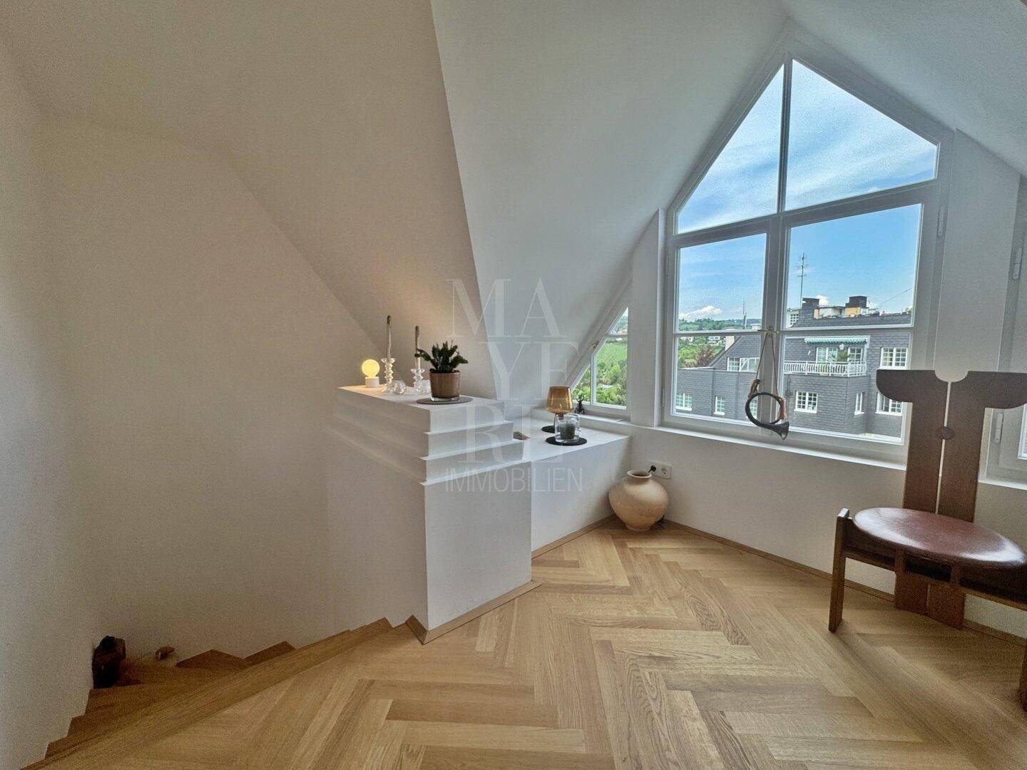 Offener Bereich mit Fischgrätparkett, Dachschräge und großem dreieckigem Fenster mit Stadtblick.