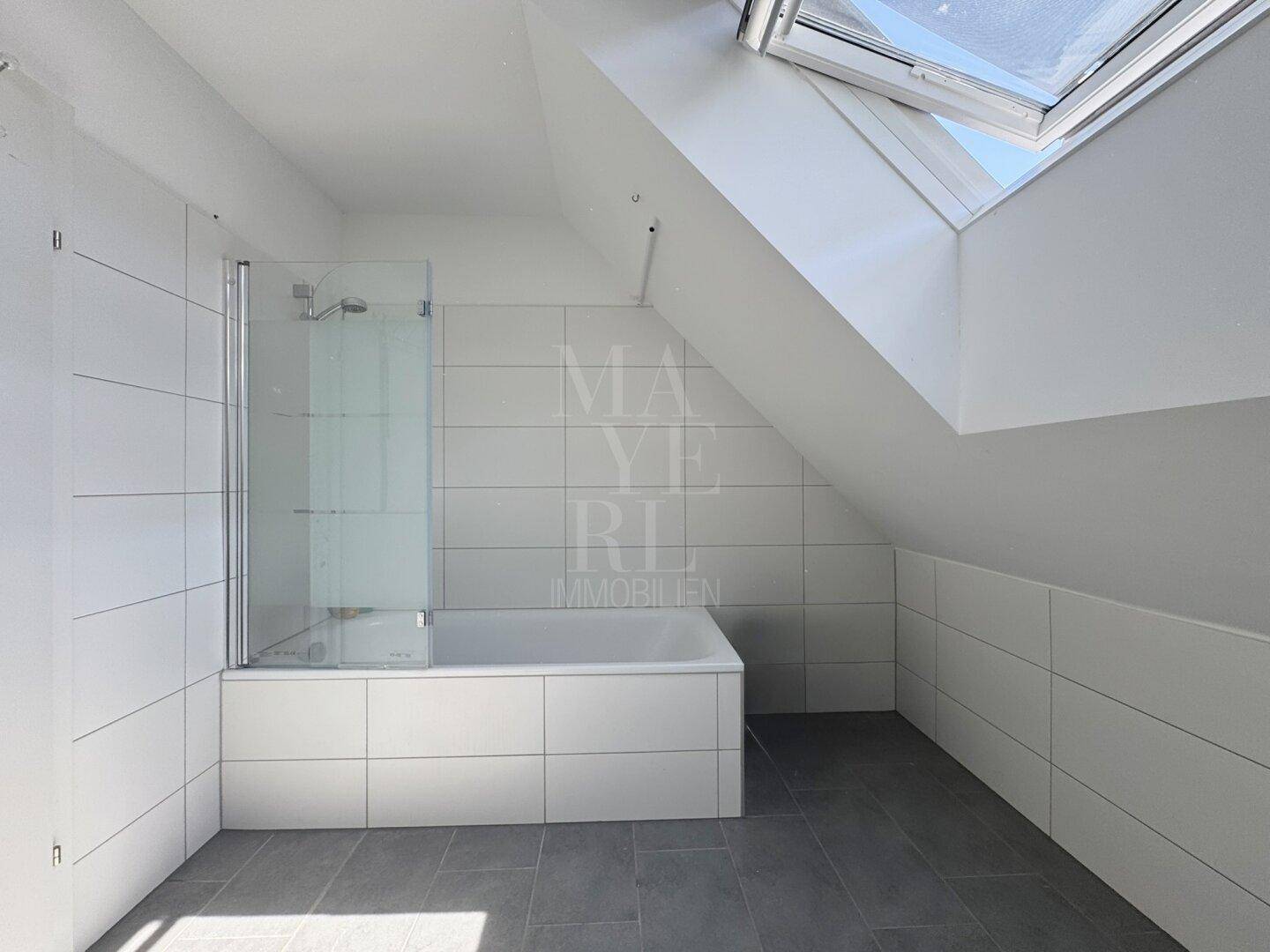 Helles Badezimmer mit Badewanne, Glasduschwand, grauen Fliesen und einem großen Dachfenster.