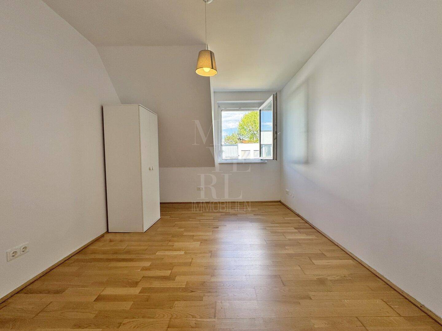 Leeres Zimmer mit hellem Holzfußboden, weißem Kleiderschrank und einem Fenster mit Blick ins Grüne.