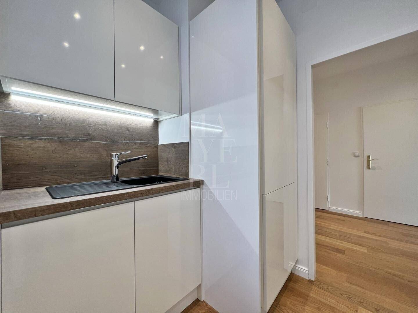 Kompakte, moderne Küche mit weißer Hochglanzfront, Spüle und integriertem Kühlschrank.