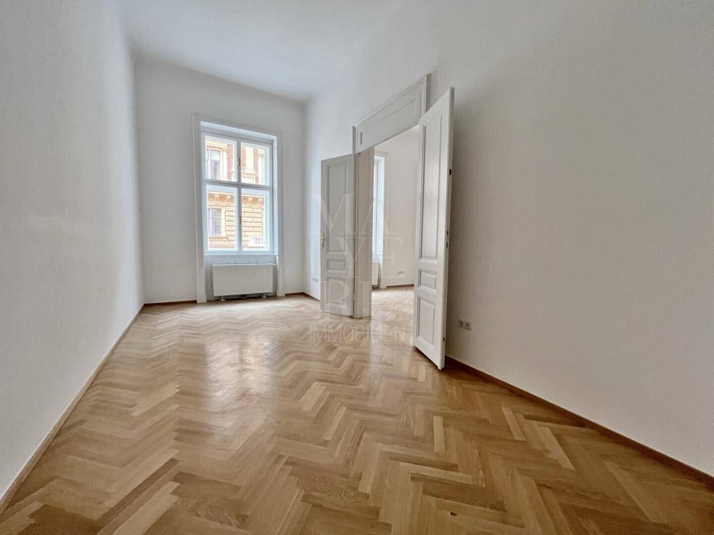 Geräumiges Zimmer mit klassischem Fischgrätparkett und hohen Doppeltüren, die zu einem weiteren Raum führen.