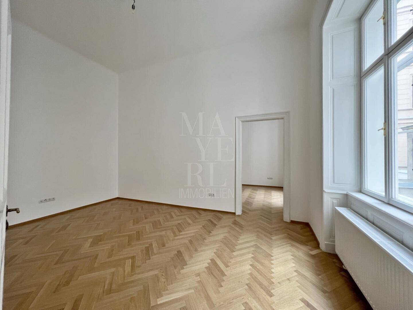 Geräumiges Zimmer mit klassischem Fischgrätparkett und hohen Decken, das eine helle und offene Atmosphäre schafft.