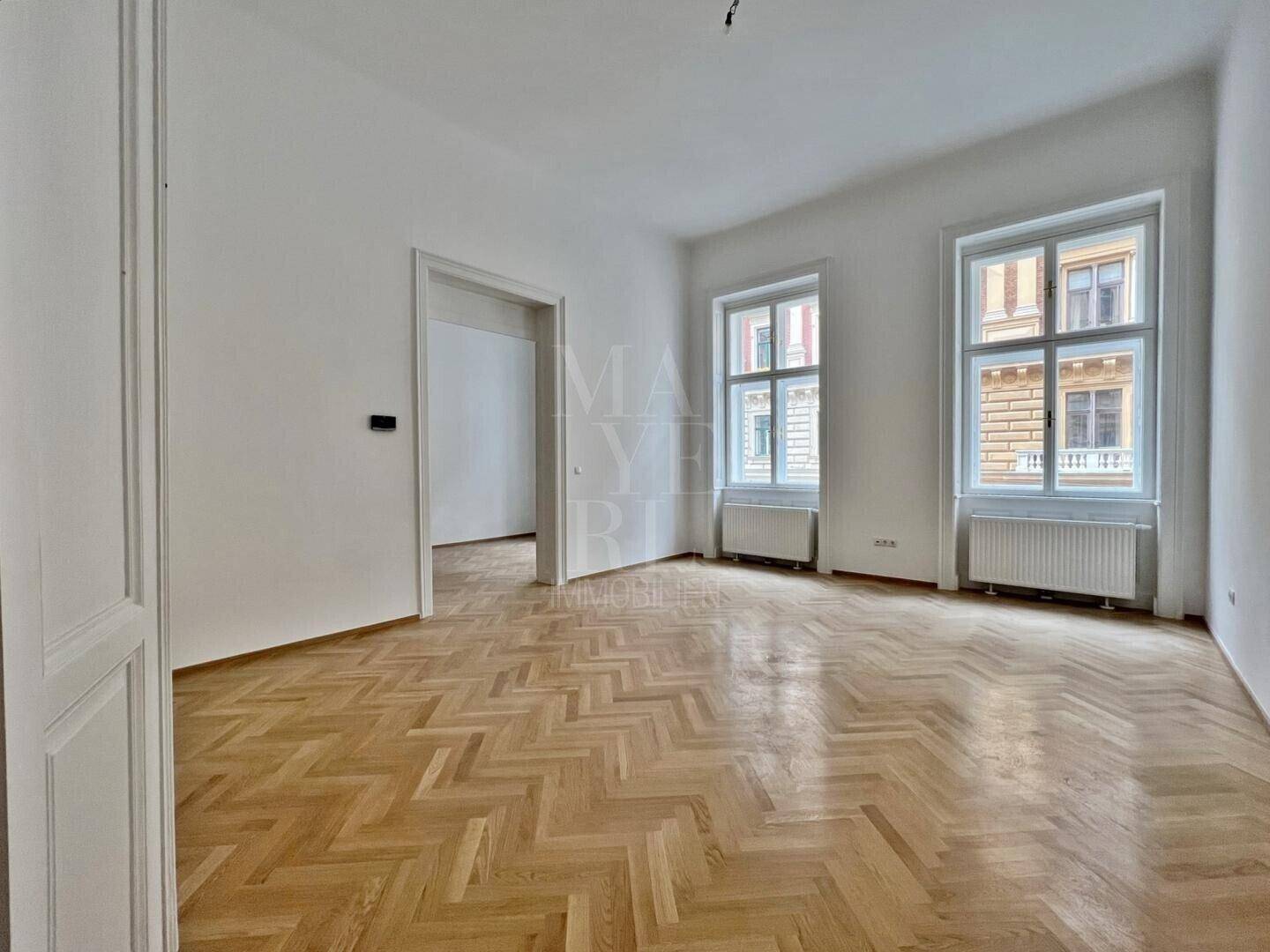 Geräumiger, heller Wohnraum mit klassischem Fischgrätparkett und zwei großen Fenstern, die viel Licht hereinlassen.