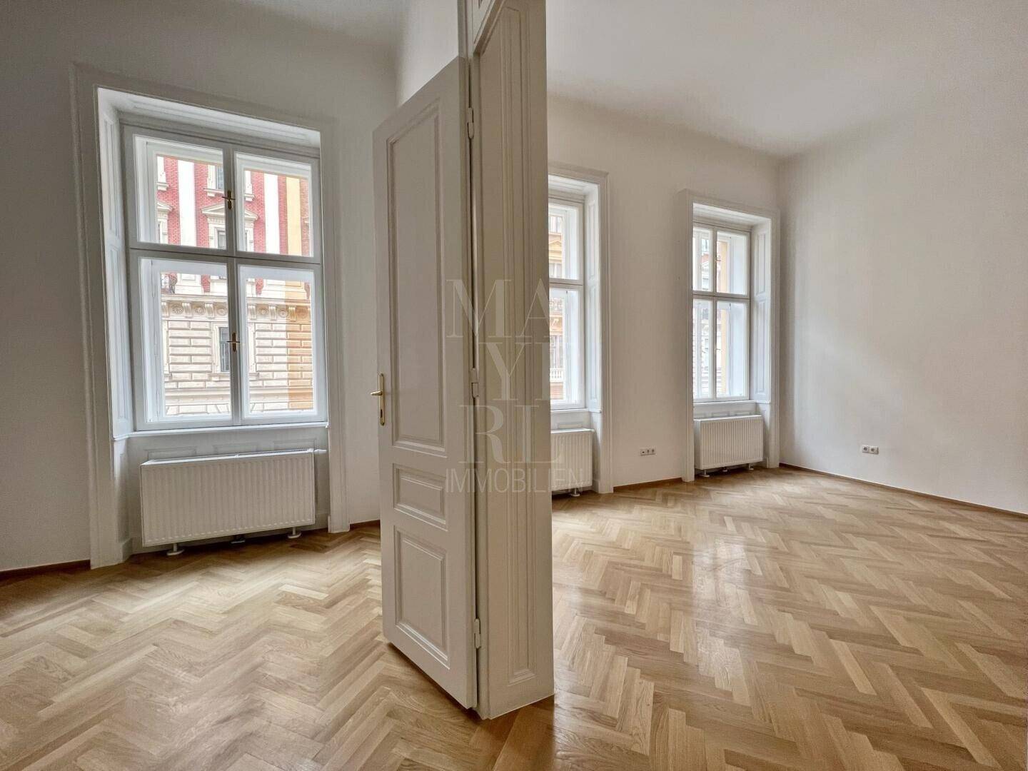 Großer, lichtdurchfluteter Raum mit Fischgrätparkett und zwei hohen Fenstern, ideal für Wohn- oder Schlafzimmer.