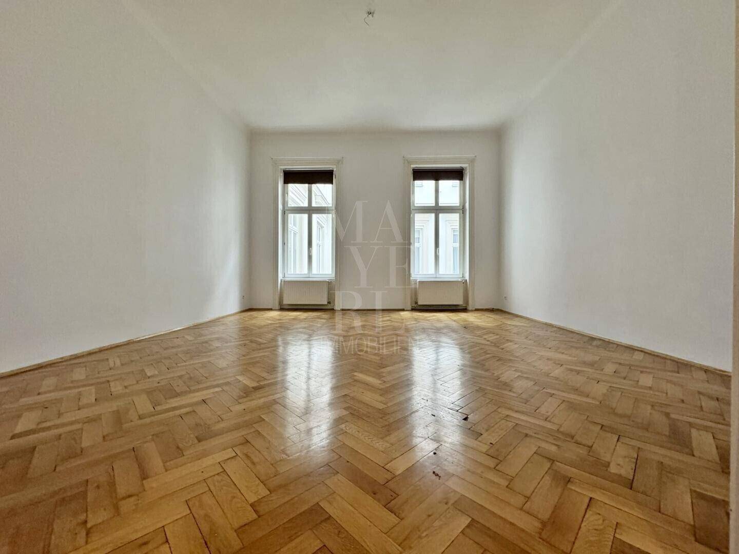 Heller, leerer Raum mit Fischgrätparkett und zwei hohen Fenstern, typisch für Altbau.