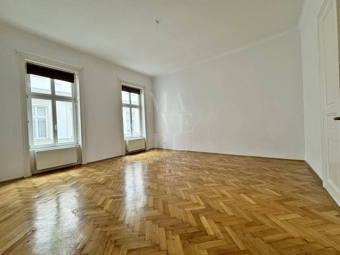 Geräumiges, leeres Wohnzimmer mit klassischem Fischgrätparkett und hohen Fenstern.