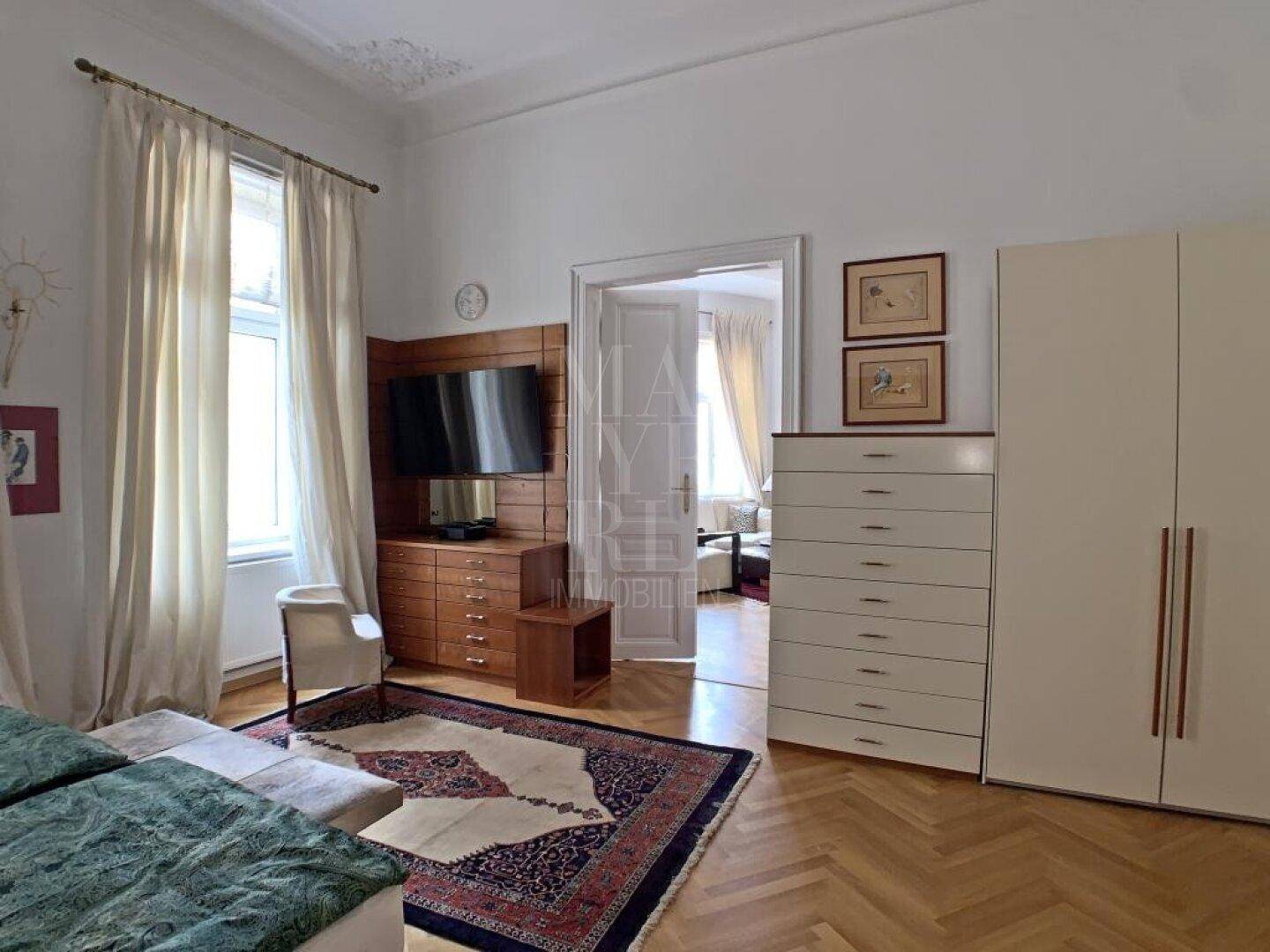 Geräumiges Schlafzimmer mit TV-Bereich, Parkettboden und Zugang zu einem weiteren Raum.