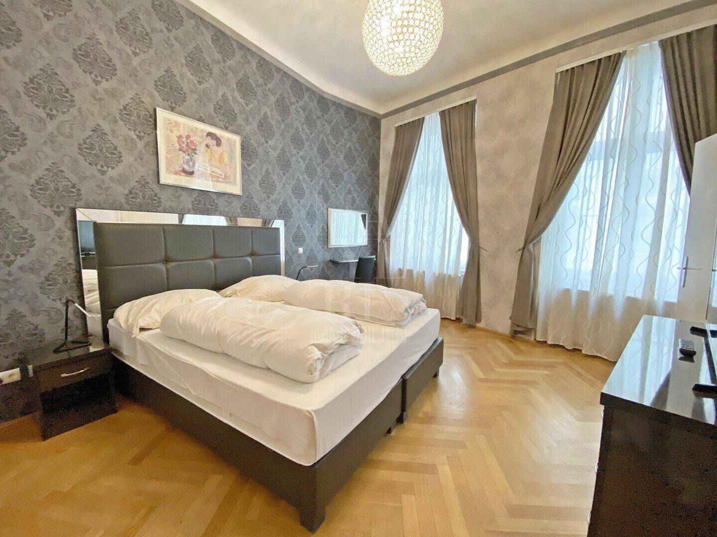 Schlafzimmer mit Doppelbett, Fischgrätparkett und großen Fenstern mit Vorhängen.